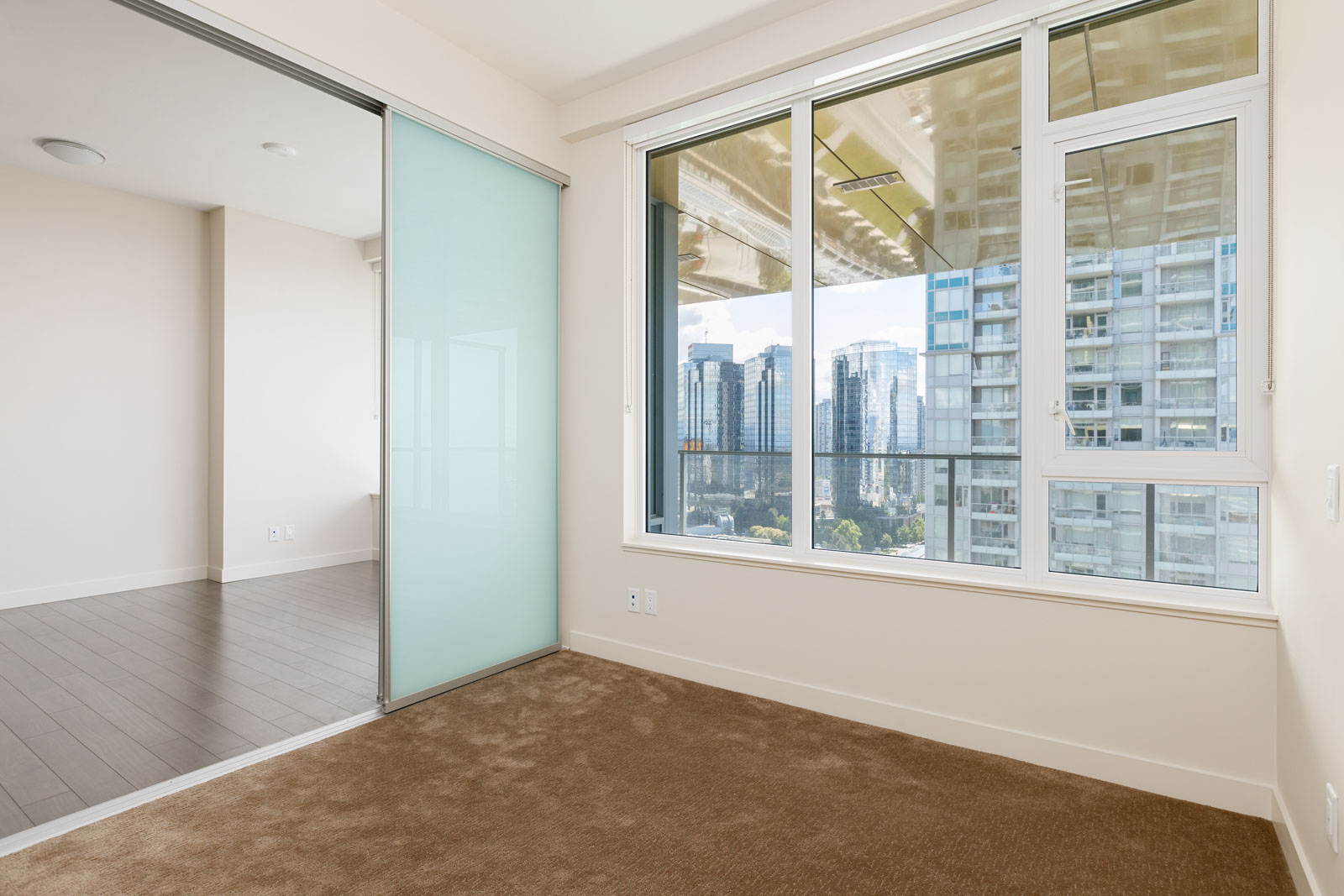 Empty room with beige carpet, glass sliding door, and large windows overlooking cityscape with high-rise buildings.