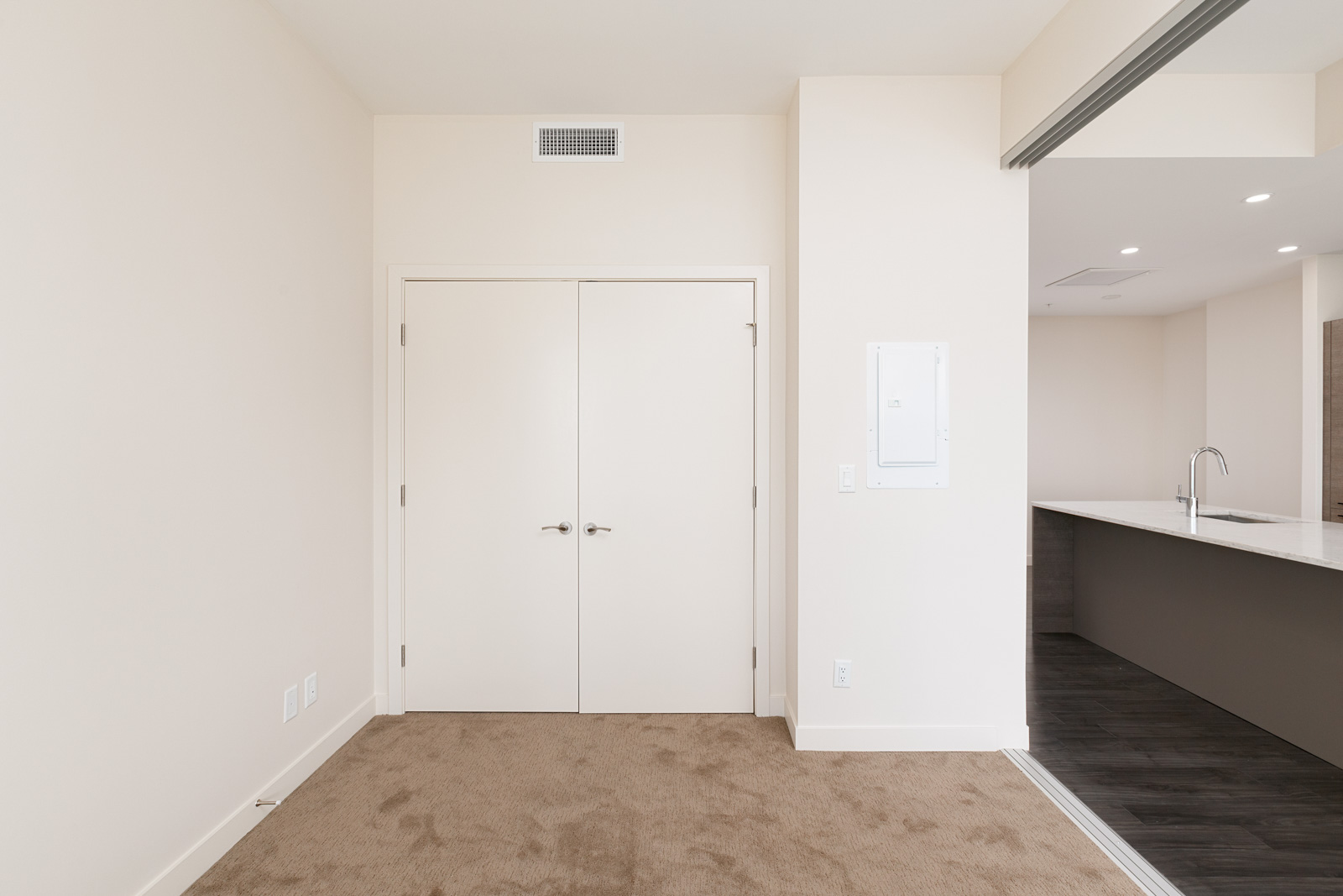 Interior view of a vacant room with beige carpet, white walls, double doors, and a partial view of a kitchen area with a countertop and sink.