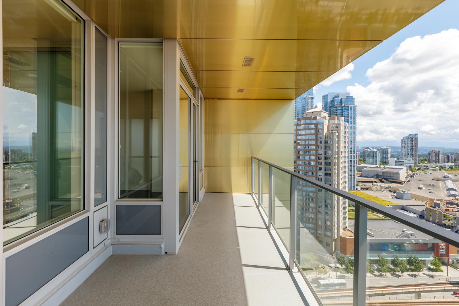 A high-rise building balcony with a glass railing offers a cityscape view of modern skyscrapers under a partly cloudy sky. The floor is light-colored, and the ceiling is a warm yellow.