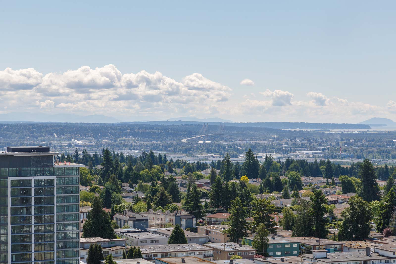 Aerial view of a cityscape with a mix of buildings, trees, and a distant highway. A high-rise building is visible on the left, under a partly cloudy sky.