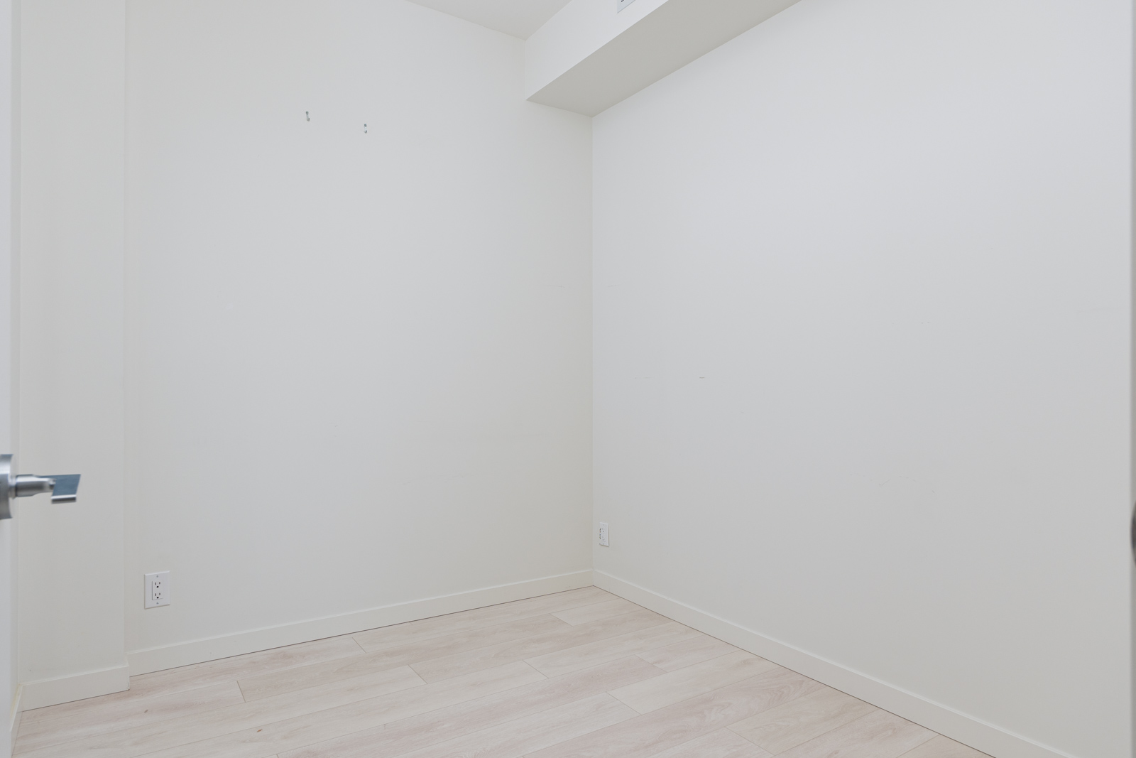 A small, empty room with white walls and light wood flooring. There's a closed door partially visible on the left. No furniture or decorations are present.