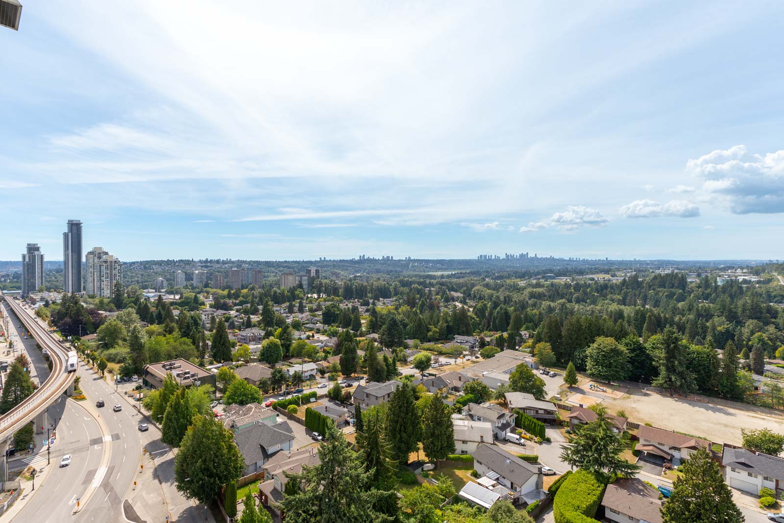 Aerial view of a suburban neighborhood with houses, roads, and trees, bordered by a highway on the left. Skyscrapers are visible in the distance under a partly cloudy sky.