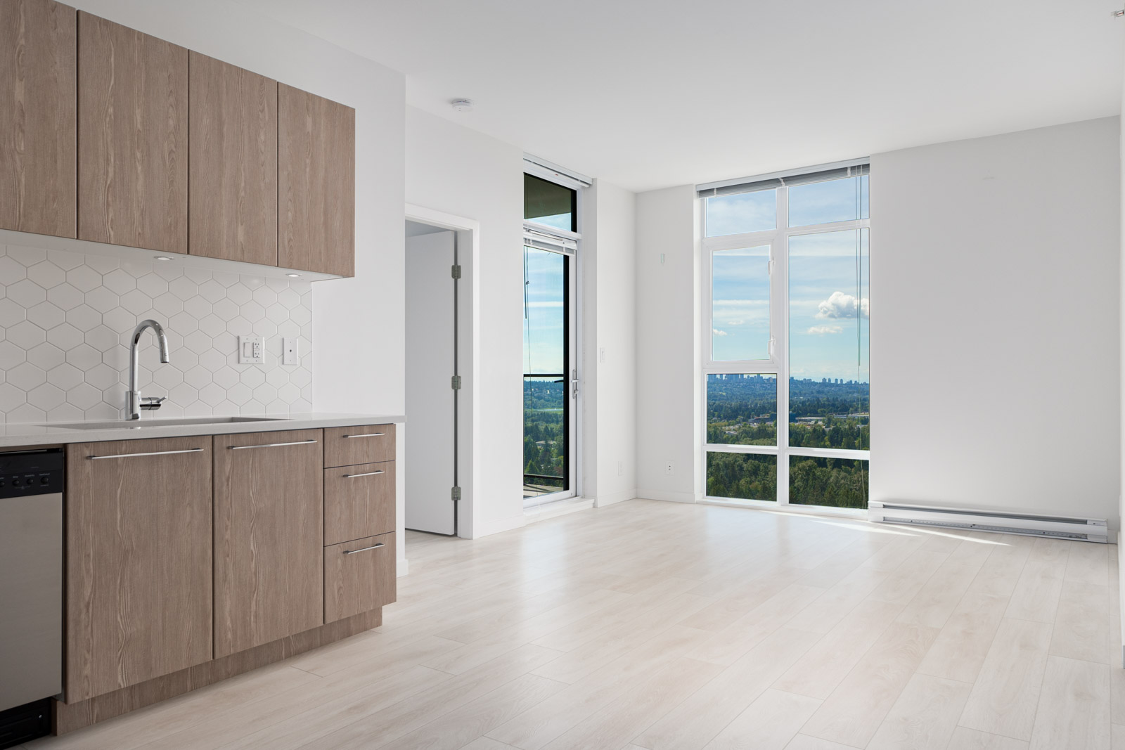 Modern empty apartment with light wood flooring, a kitchen area on the left featuring wooden cabinets, and large windows on the right offering a clear view of the outdoors.