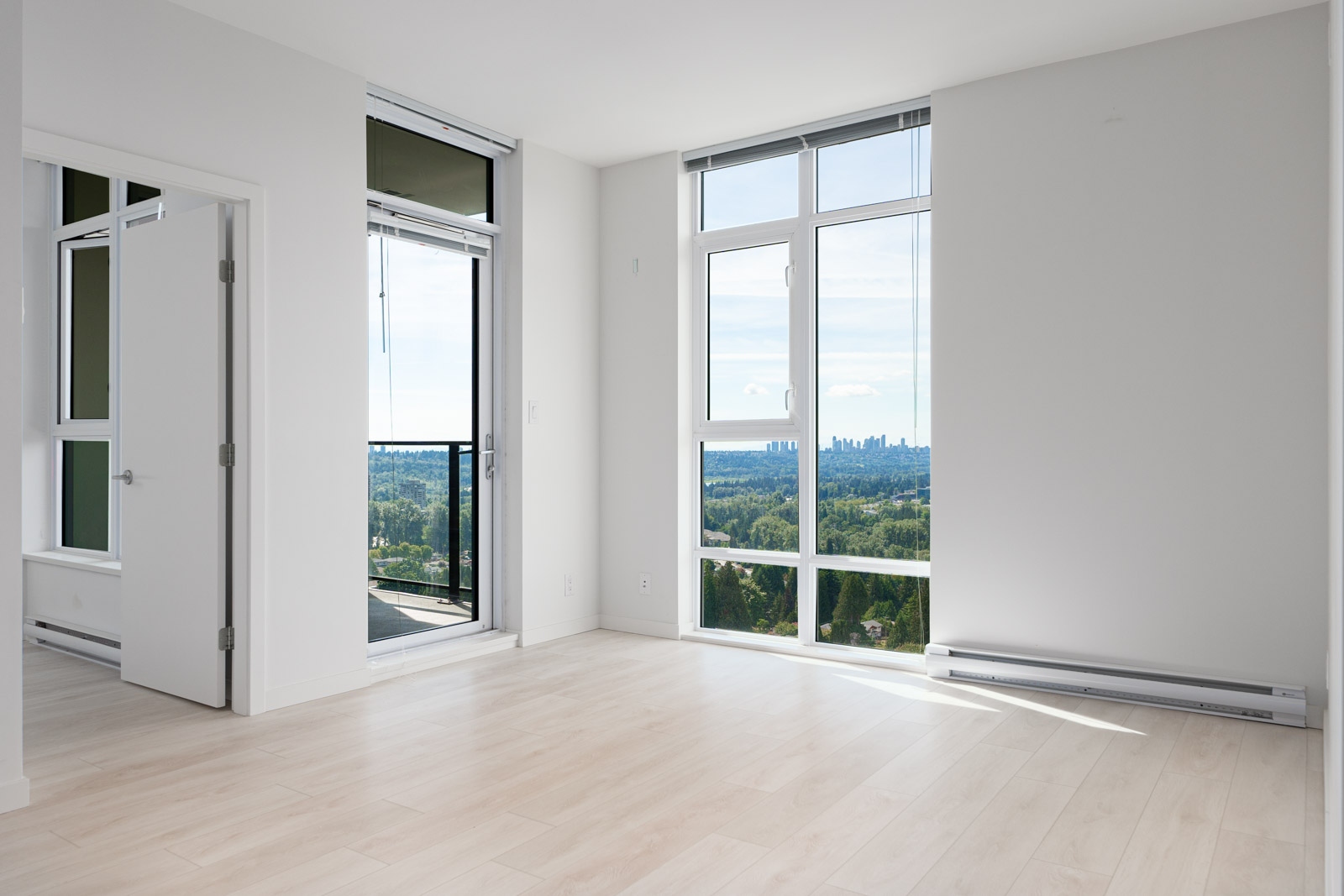 Bright, empty room with light wood flooring, large windows, and a balcony offering a view of a city skyline in the distance.