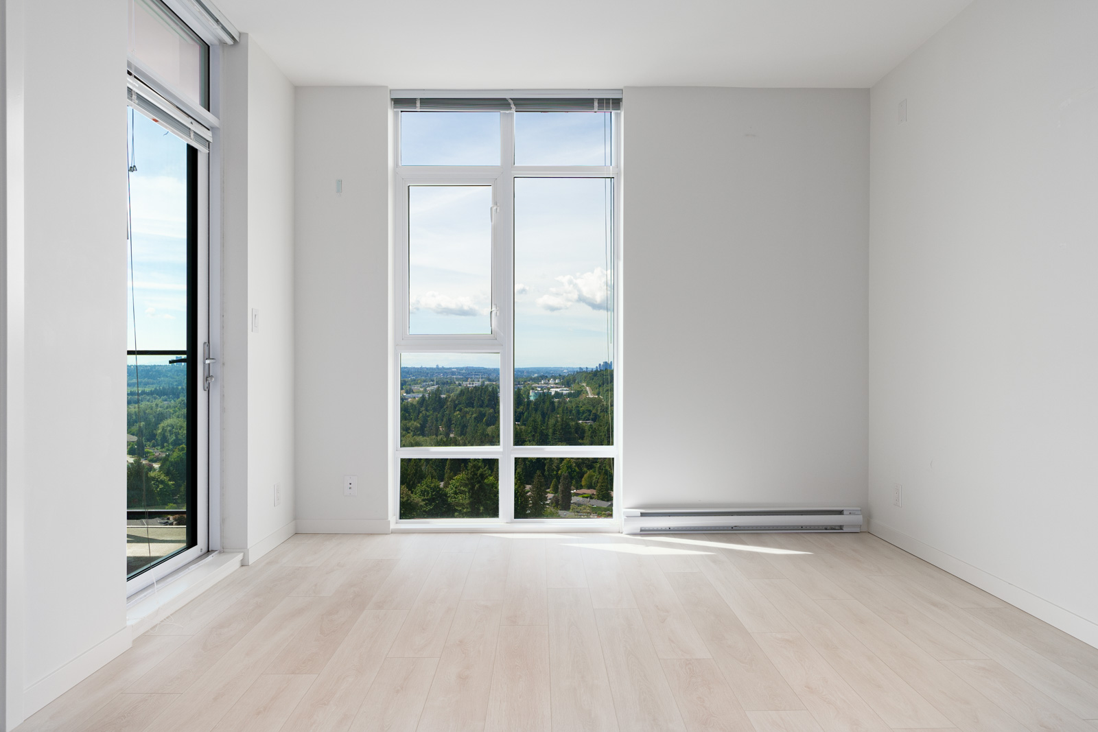 An empty room with light wood flooring, large windows, and a door leading to a balcony. The windows offer a view of greenery and a distant body of water under a partly cloudy sky.