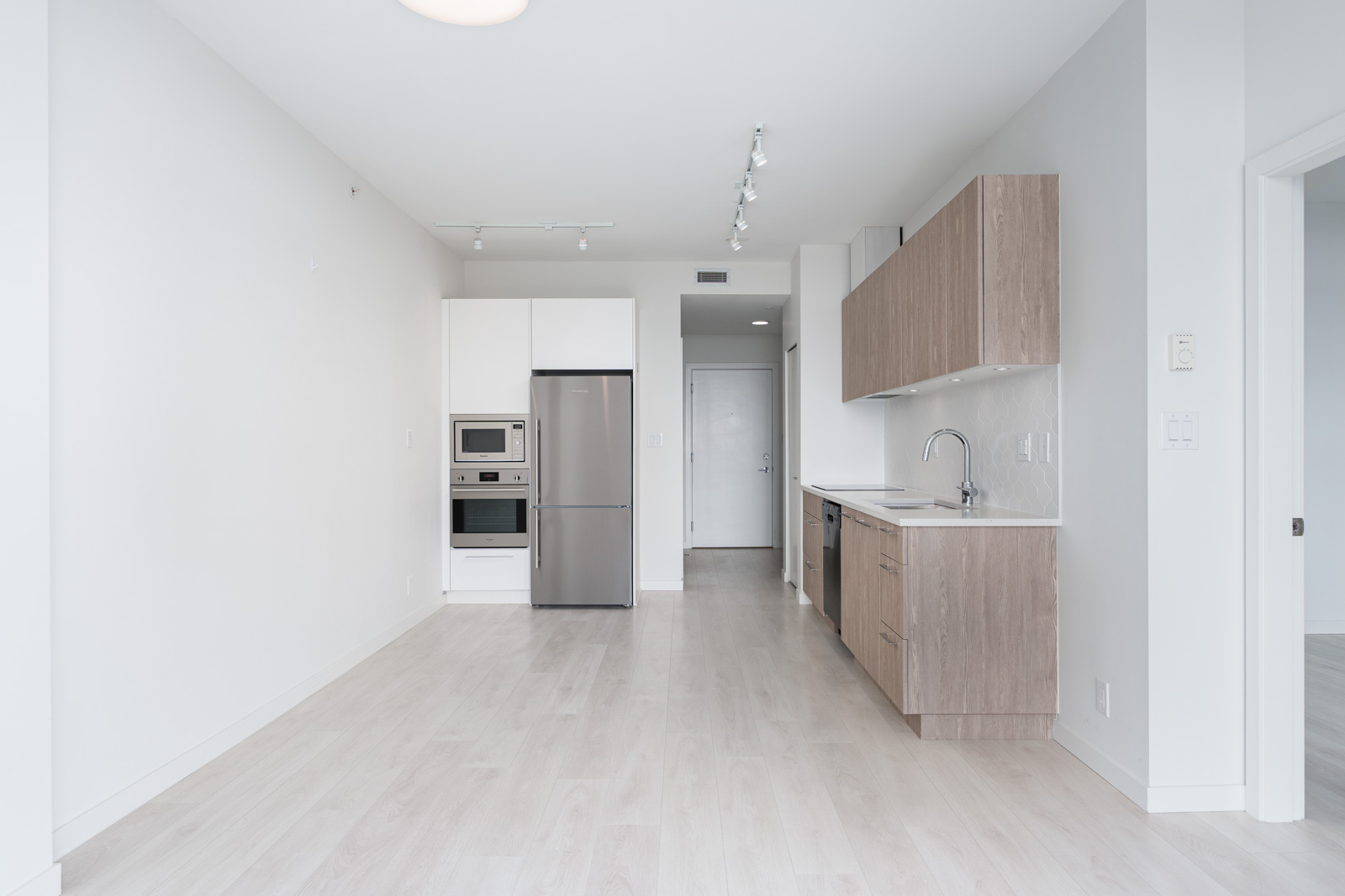 Modern kitchen with stainless steel appliances, light wood cabinets, and light wood flooring, leading to a hallway with a white door.