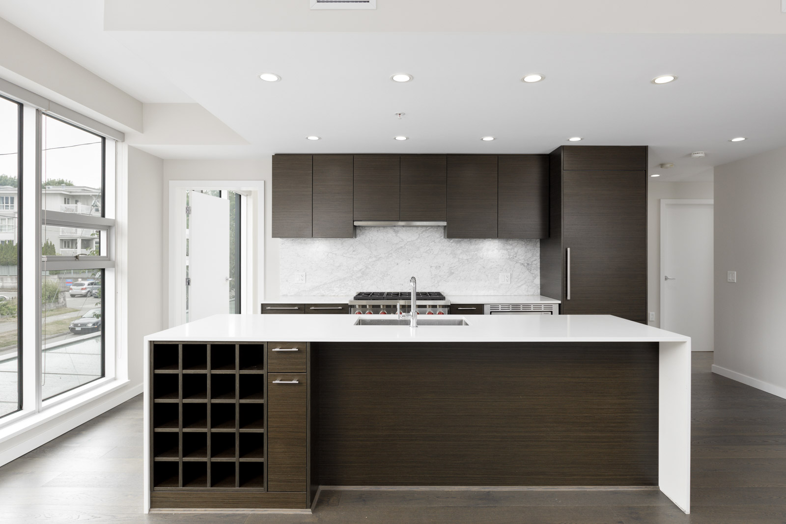 Modern kitchen with dark wood cabinets, a white island with built-in wine rack, stainless steel appliances, and large windows providing natural light.