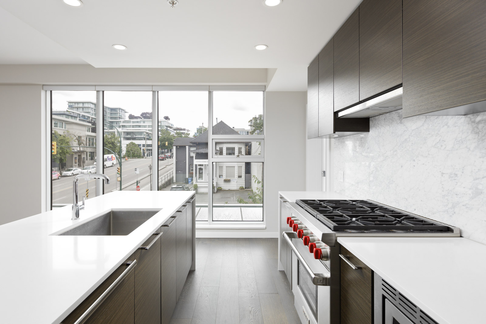 Modern kitchen with white countertops, stainless steel appliances, and large windows overlooking a street and residential buildings.
