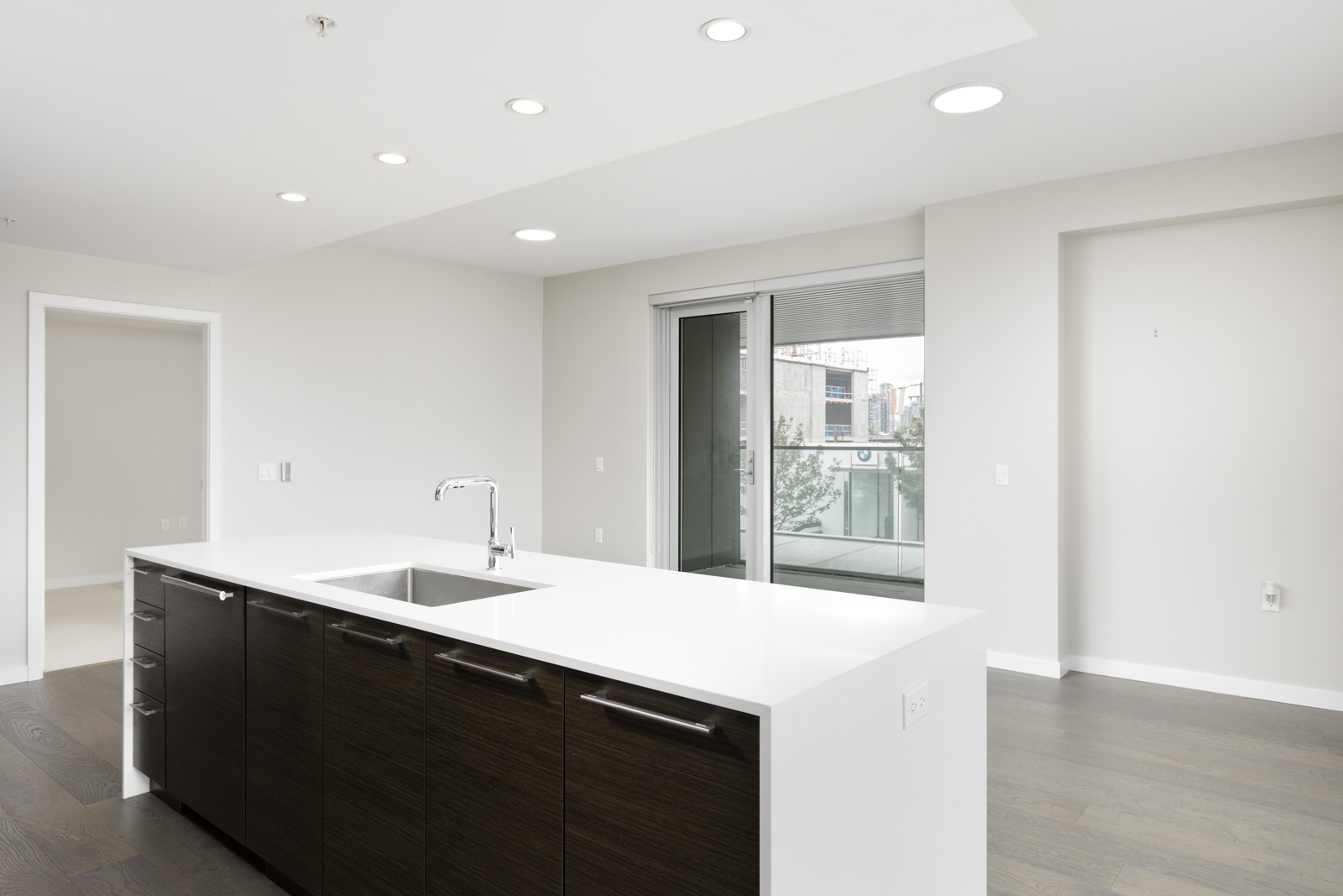 Modern kitchen with a white island, dark wood cabinets, built-in sink, and sliding glass doors opening to a balcony in a bright, unfurnished apartment.