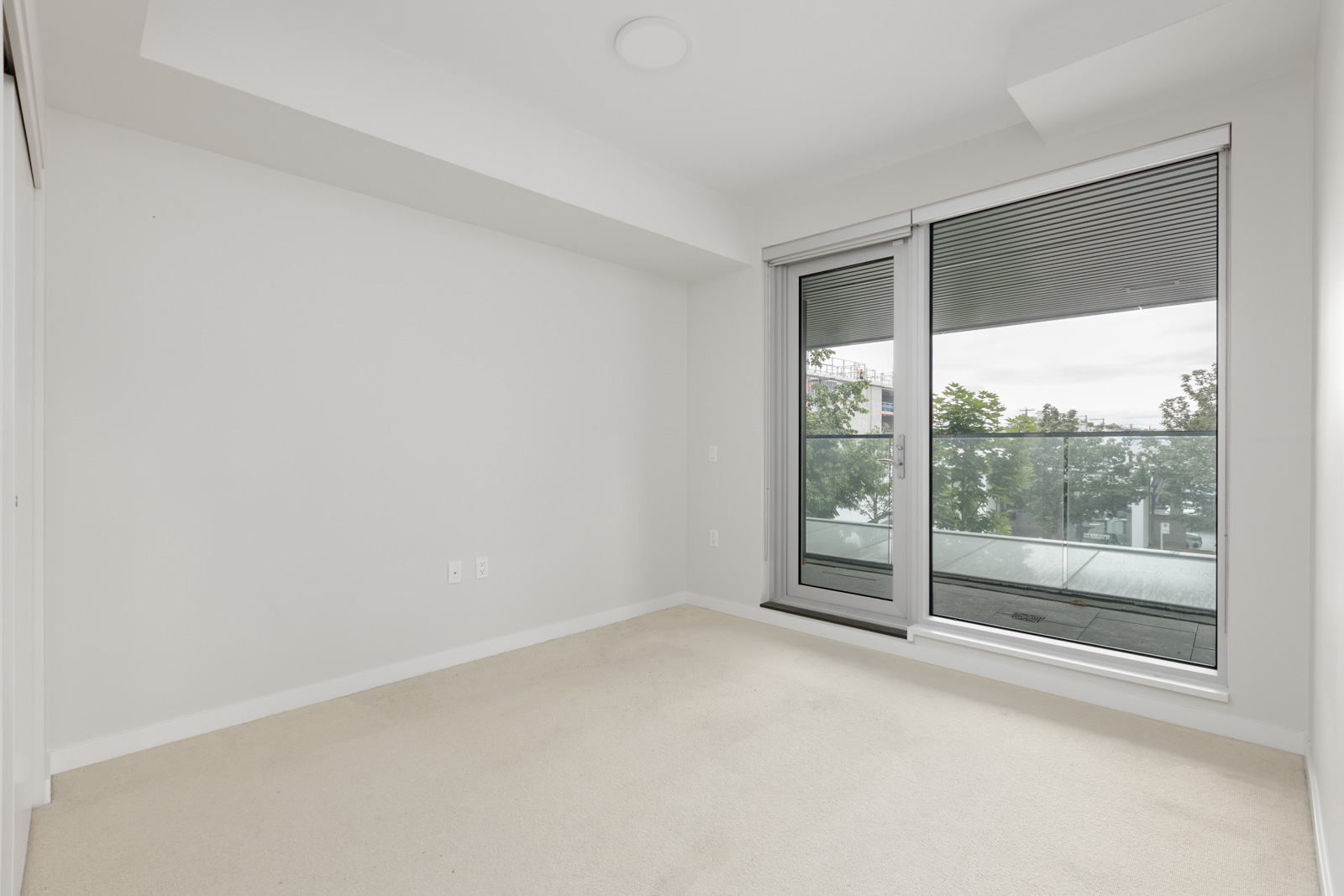 Empty room with light-colored carpet, white walls, and large glass door leading to a balcony with trees visible outside.