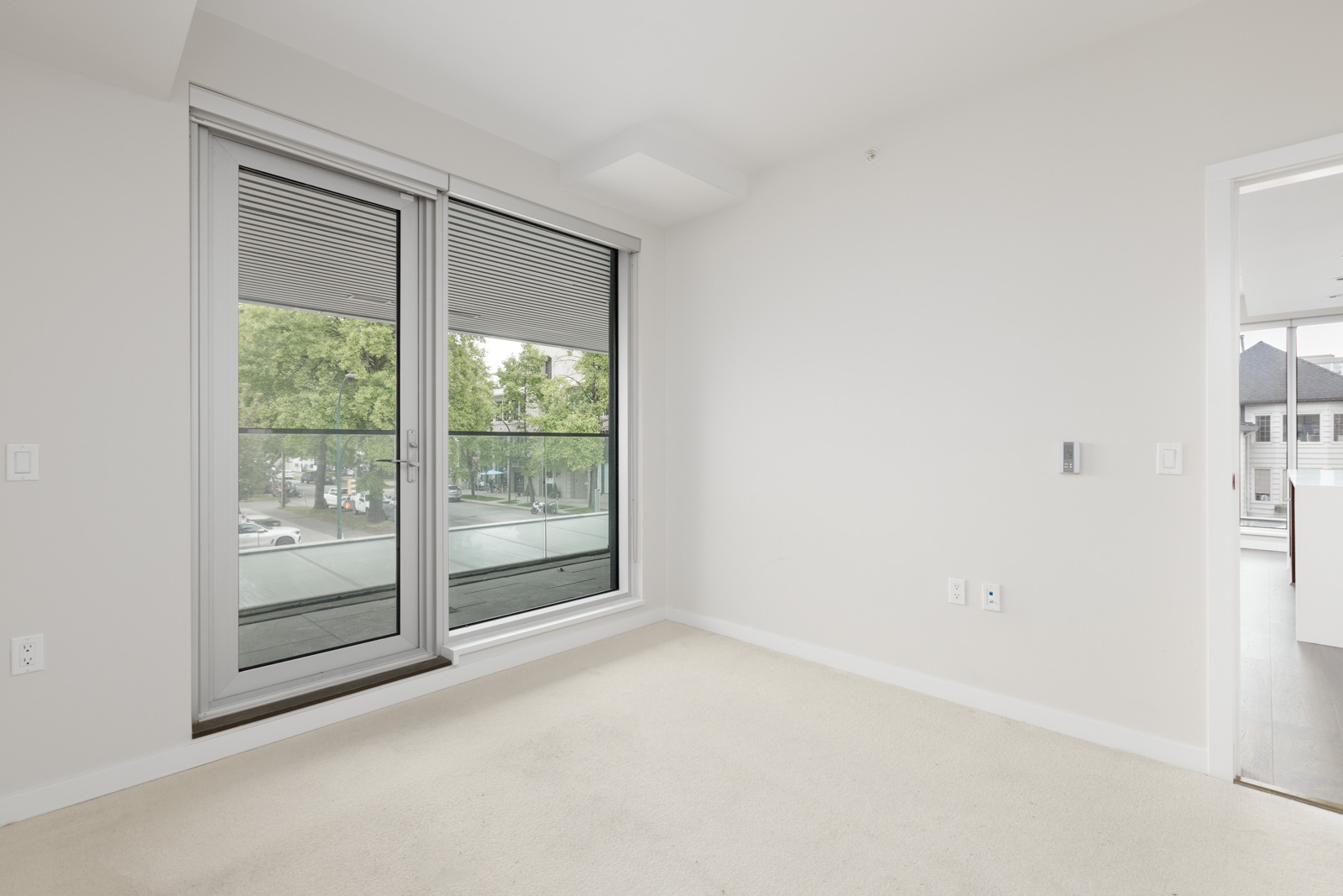 Empty bedroom with beige carpet, large glass doors leading to a balcony, and a view of trees and parked cars outside. An open doorway leads to another room.