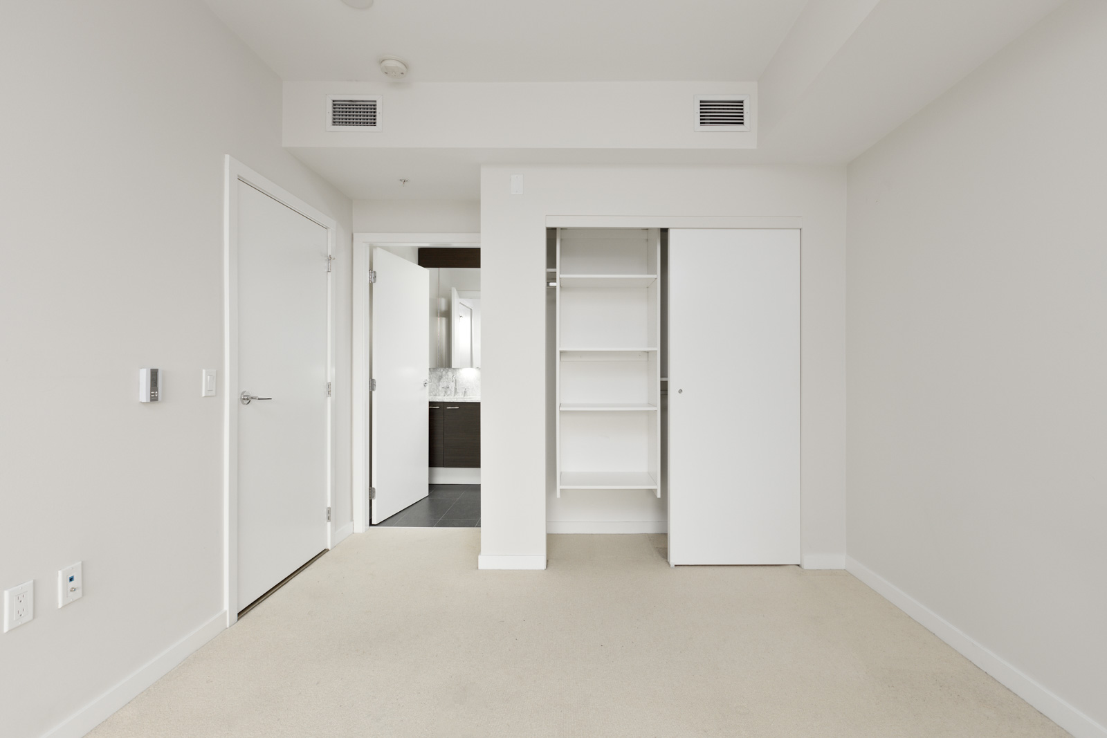 A clean, empty bedroom with beige carpet, white walls, an open closet with shelves, a closed door, and a view into a bathroom.