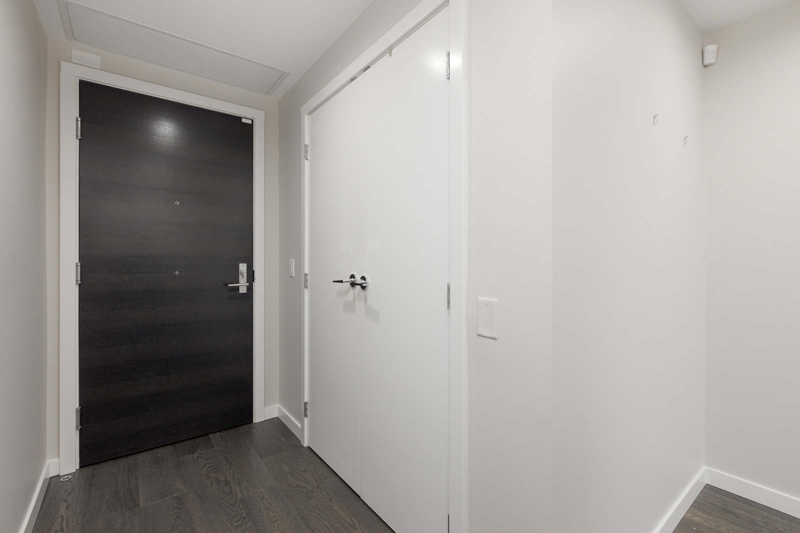 A modern entryway with a dark wooden door, white walls, double white closet doors, and dark hardwood flooring.