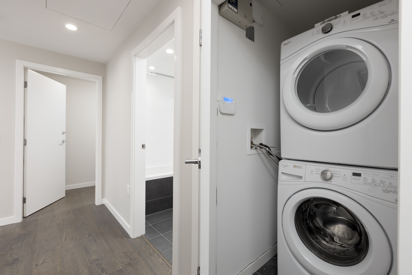 A small laundry area with a stacked washer and dryer is next to a hallway and a bathroom with a bathtub.