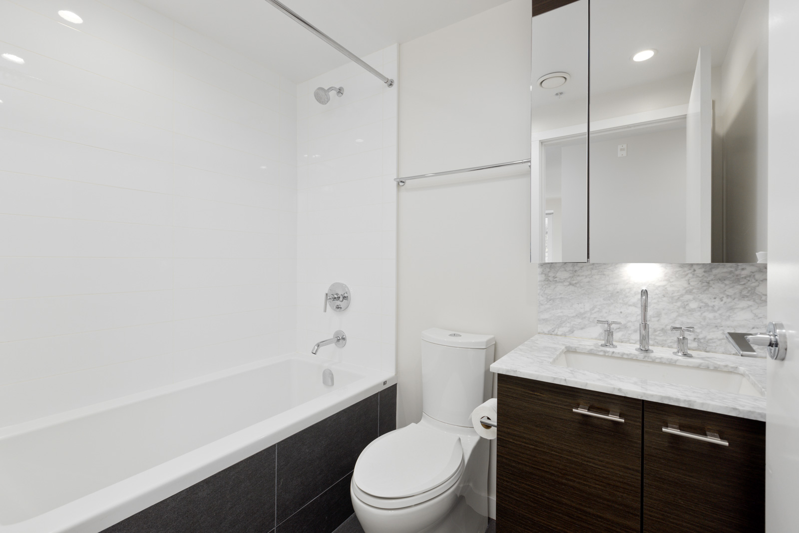Modern bathroom with a bathtub, shower, toilet, and a vanity with a marble countertop and sink. The space features white walls and dark wood cabinetry.