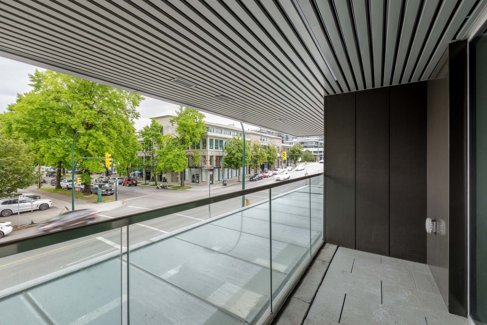 View from a covered balcony with glass railing overlooking a city street, green trees, and buildings on a cloudy day.