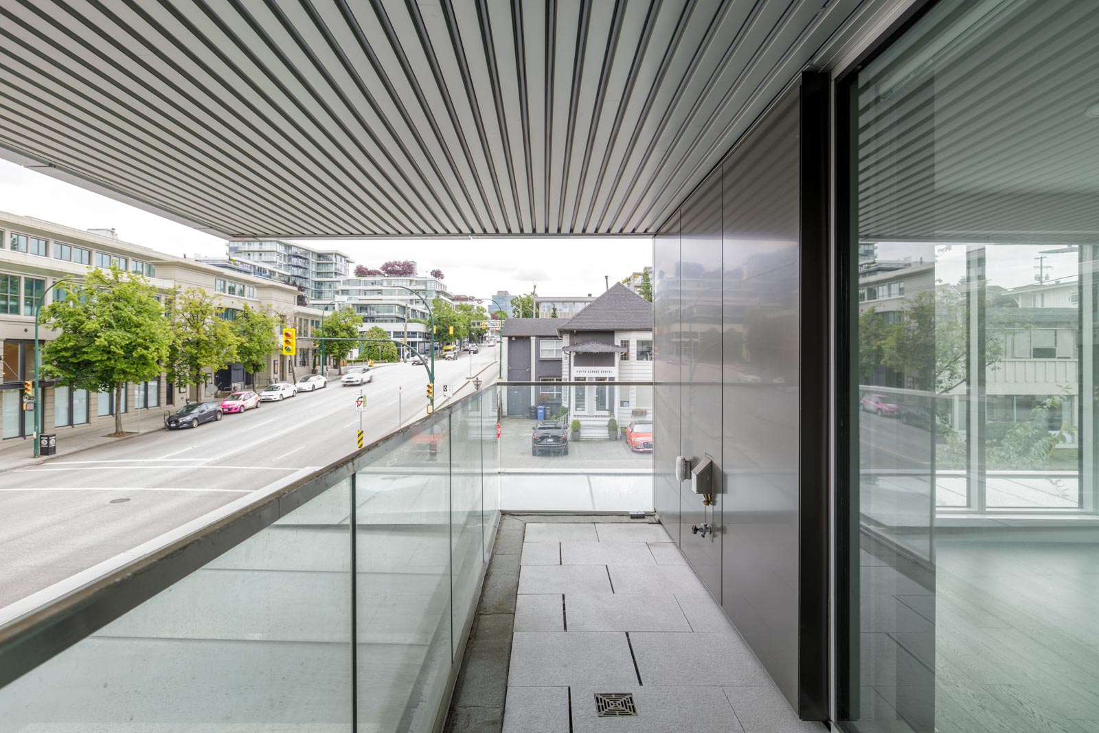 A modern glass balcony overlooks a quiet urban street lined with trees, parked cars, and contemporary buildings on a cloudy day.
