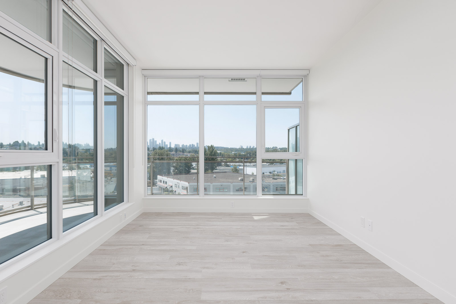 Bright, empty room with large windows, light wood flooring, and a view of the city skyline in the distance.