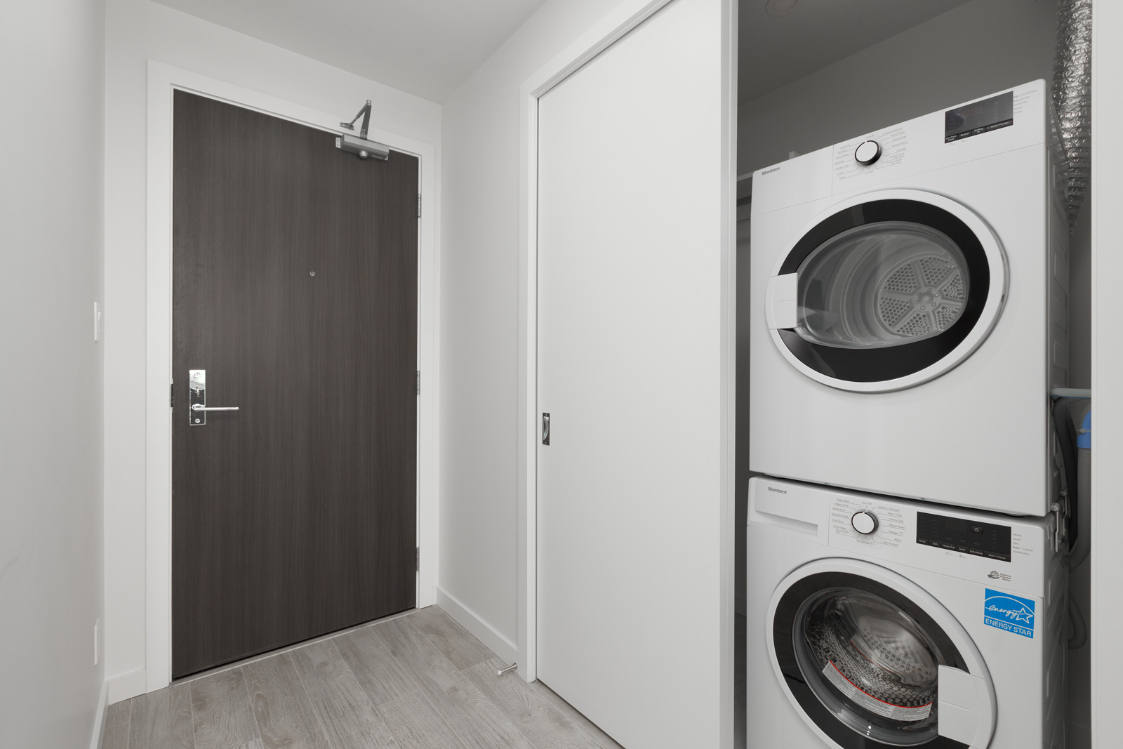 Laundry area with a stacked washer and dryer next to a closed wooden door and a sliding white closet door.