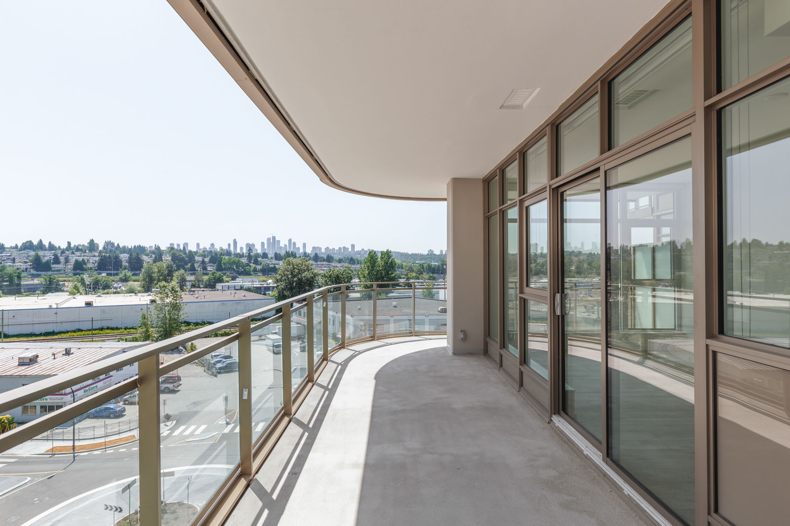 Spacious curved balcony with glass railing, overlooking a cityscape and distant skyline. Large windows line the adjacent building wall.