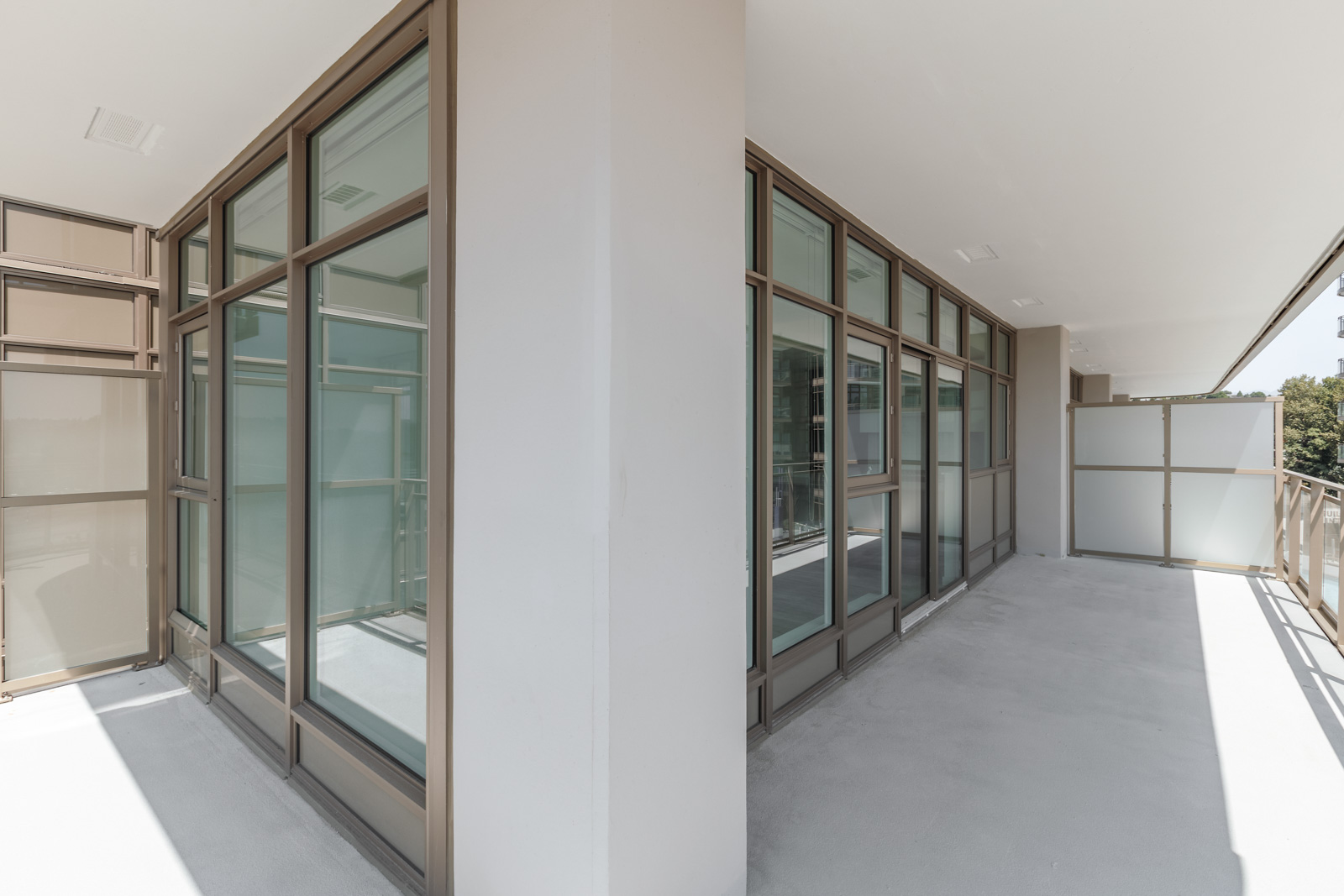 A wraparound balcony with glass and metal railings and large windows. The balcony features light-colored flooring and a dividing column.