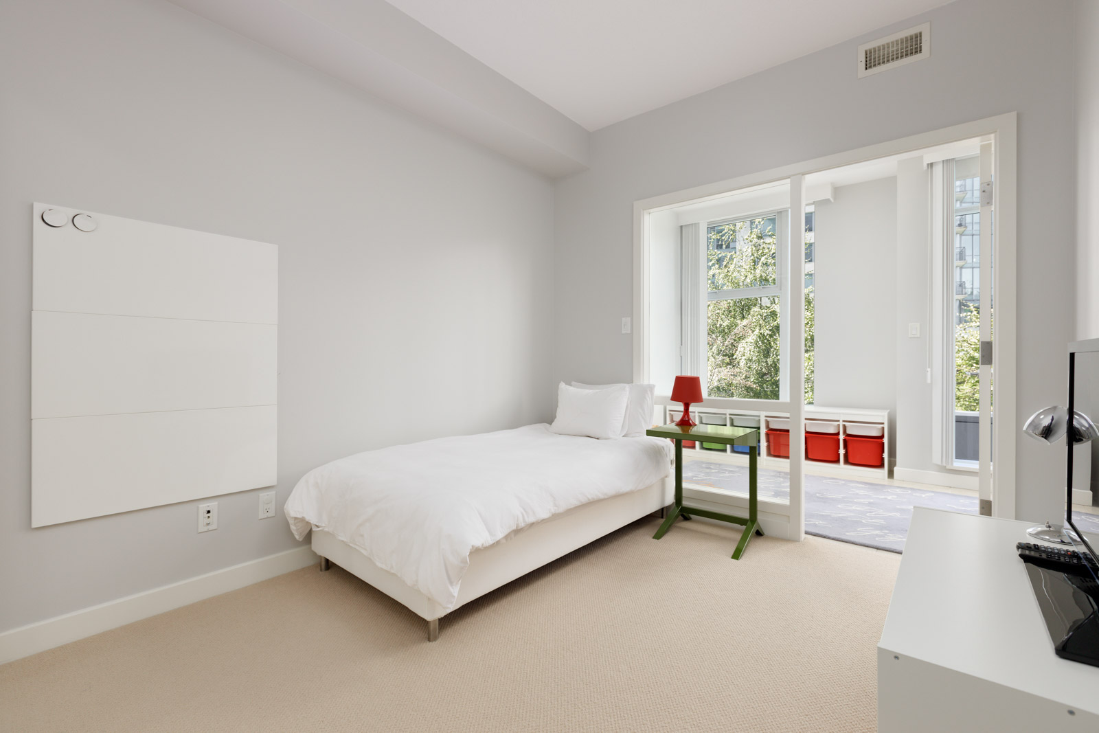 Minimalist bedroom with a single bed, white bedding, small green desk, and a sliding glass door opening to a bright sunroom with red storage bins.
