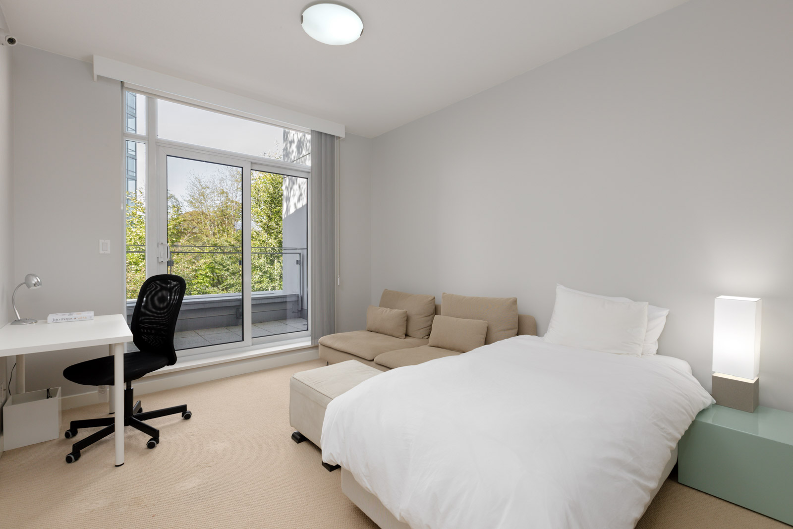 A modern bedroom with a single bed, beige sofa, white desk and office chair, large window, and glass door opening to a balcony with trees visible outside.