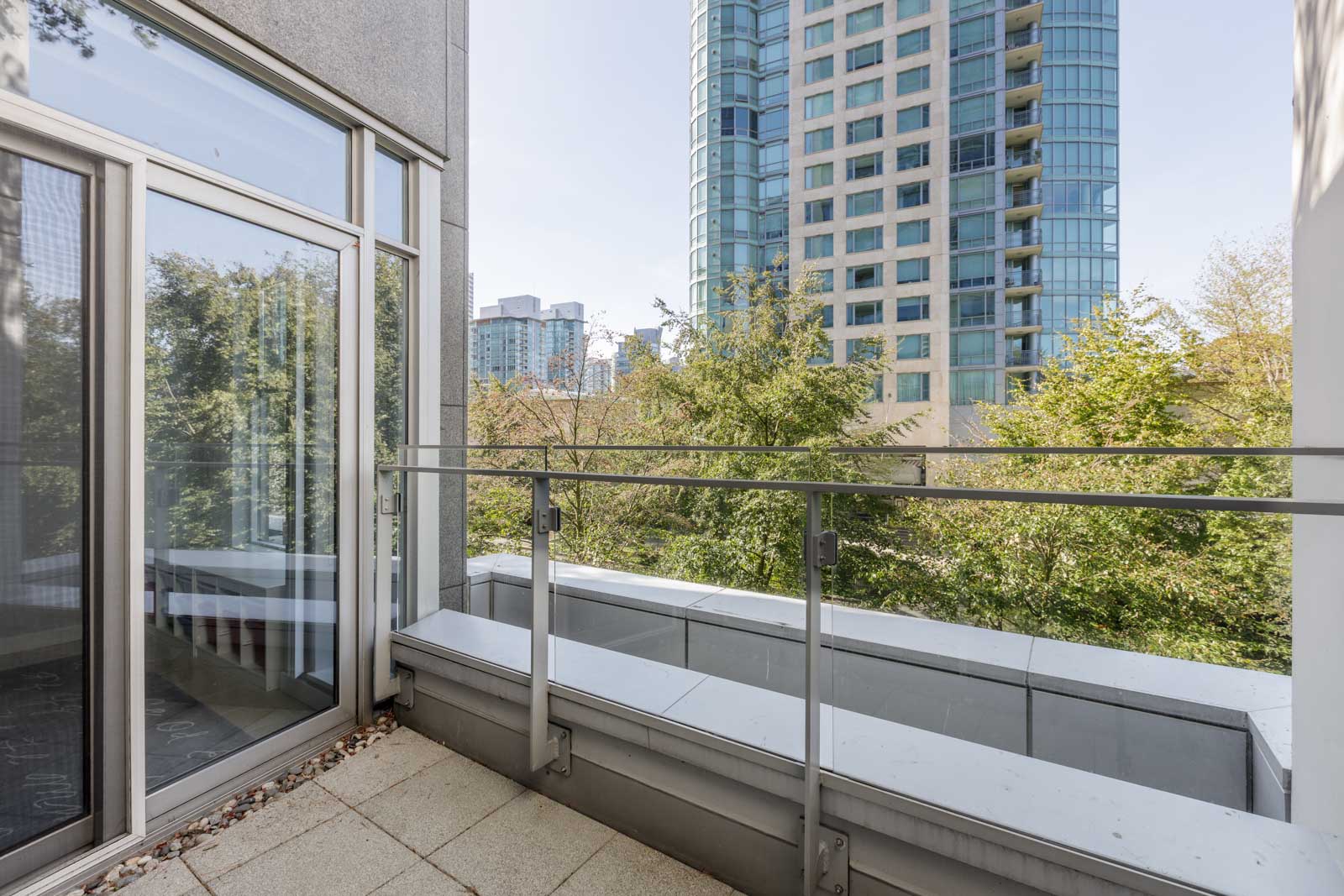 A modern balcony with glass railing overlooking trees and a tall residential building in the background on a clear day.