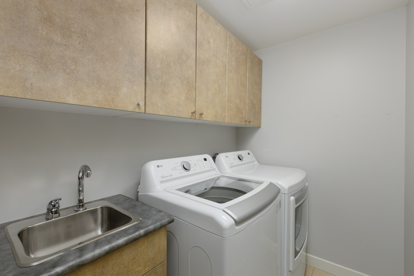 Laundry room with a stainless steel sink, upper wooden cabinets, a washing machine with the lid open, and a front-loading dryer against a plain white wall.