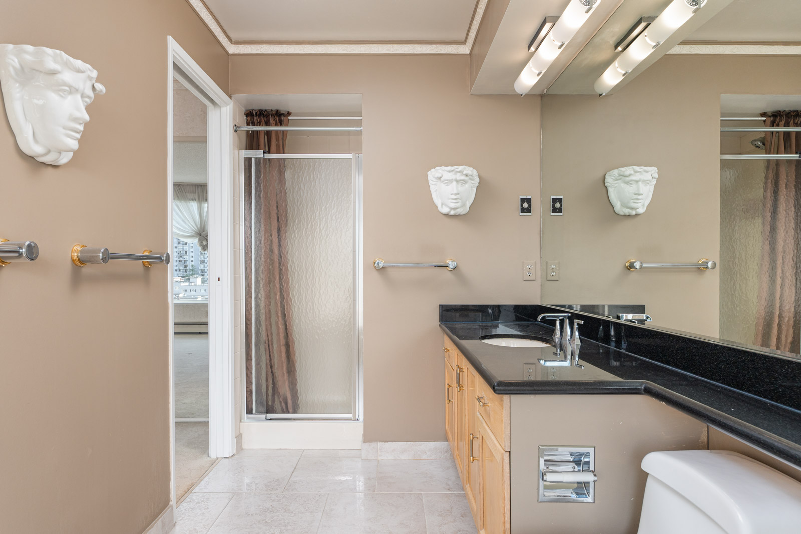 Bathroom with a large mirror, black countertop, and wooden cabinets. Wall art of faces is above towel racks. Semi-transparent glass door leads to a separate room. Ceiling has strip lights.