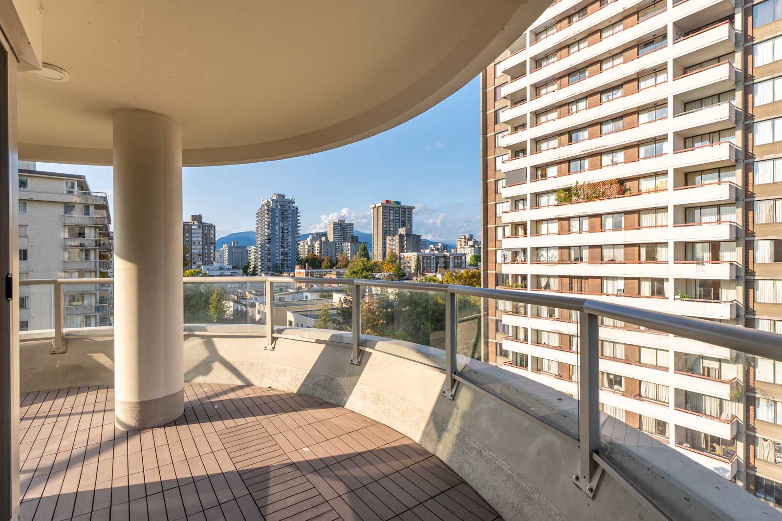 A curved balcony with wooden flooring and a glass railing overlooks a cityscape with modern buildings under a clear sky.