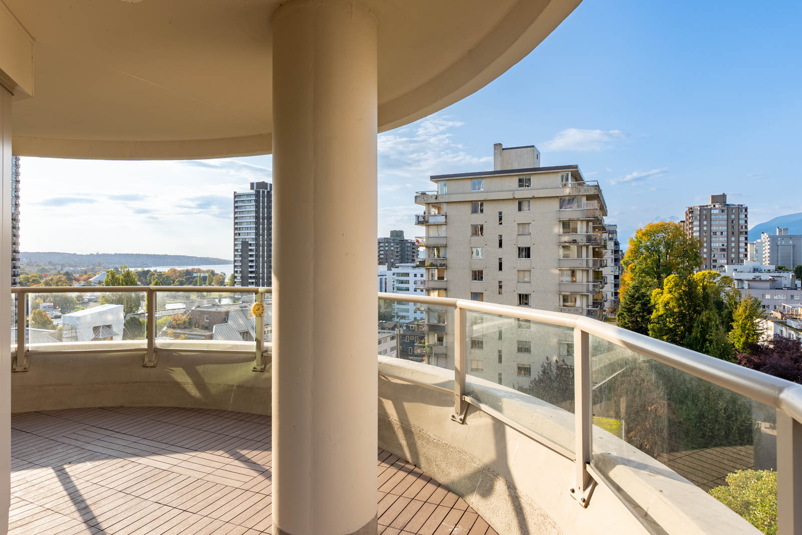 Curved balcony with glass railing, offering a city view featuring mid-rise buildings and trees under a clear sky.