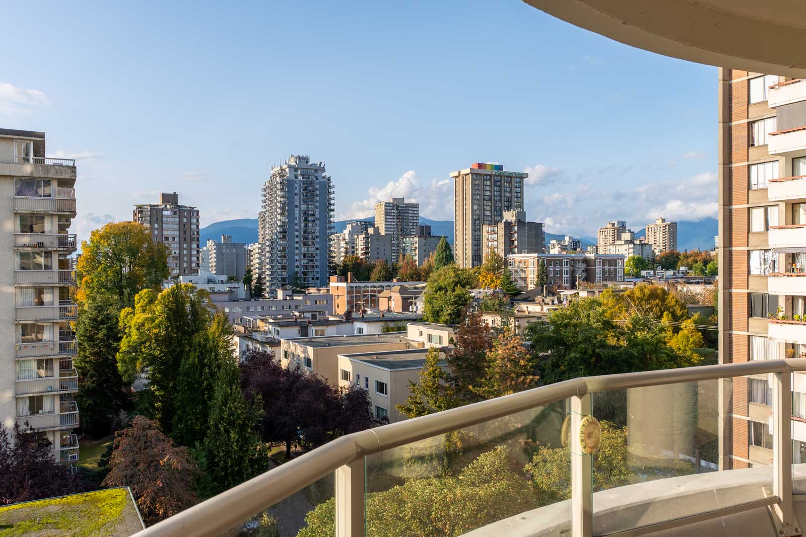 Cityscape view from a balcony overlooking buildings, trees, and distant mountains under a clear blue sky.