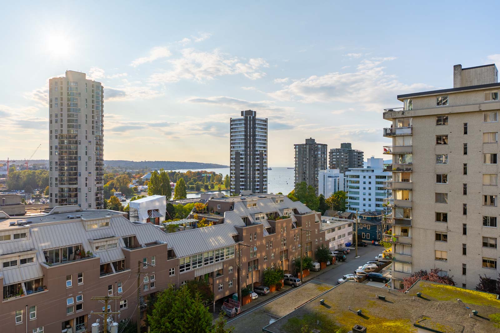 Cityscape with residential buildings under a clear sky. Sunlight reflects off the tallest structure. Trees and a glimpse of water are visible in the background.
