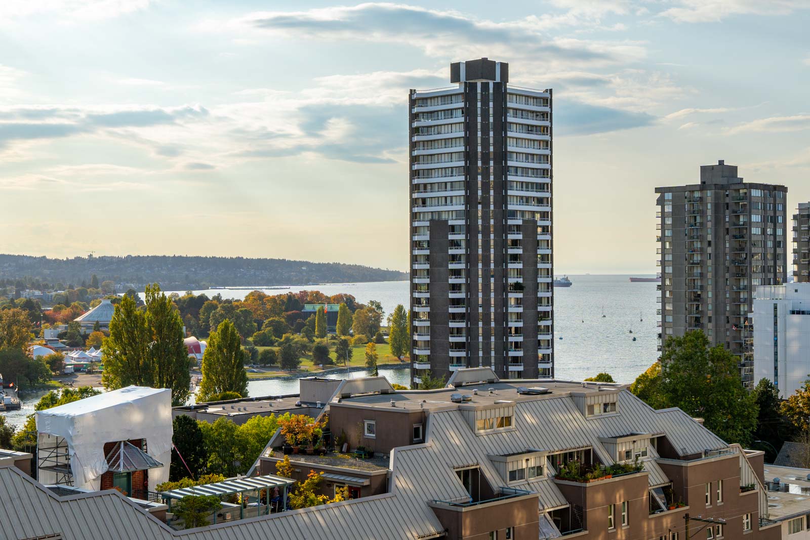 Urban landscape with tall buildings near a waterfront. Trees and a park are visible in the foreground, with a body of water and ships in the background under a partly cloudy sky.