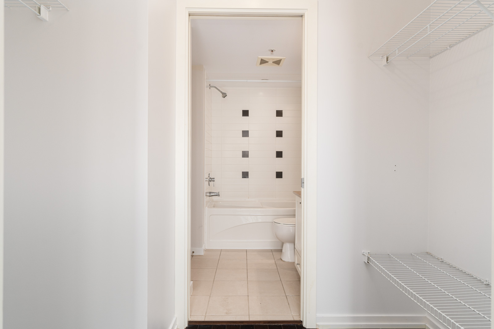 A view of a bathroom with a bathtub and shower, white tiled walls with black accent tiles, a toilet, and a metal shelving unit in the adjacent space.
