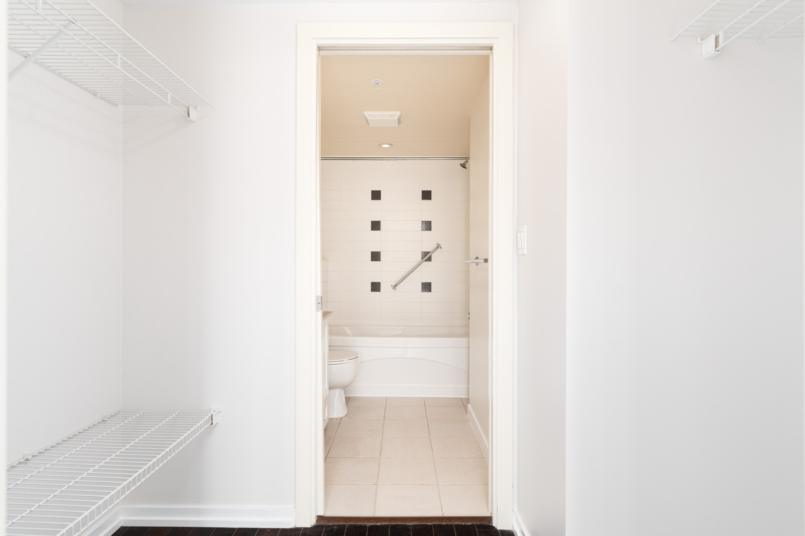 View through an open doorway into a bathroom with a bathtub, white tile walls, black square accents, and a grab bar; wire shelving is visible on the left.