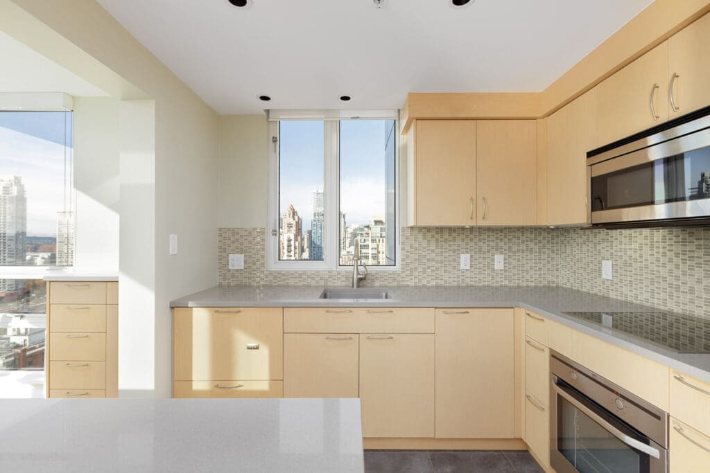 Modern kitchen with light wood cabinets, gray countertops, built-in oven, and a city view through the window. Neutral-toned backsplash and tiled floor complement the design.