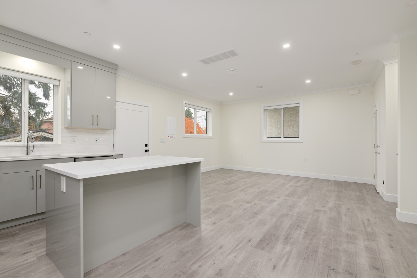 Modern, unfurnished open-plan kitchen and living area with light gray flooring, white walls, large windows, and recessed ceiling lights.