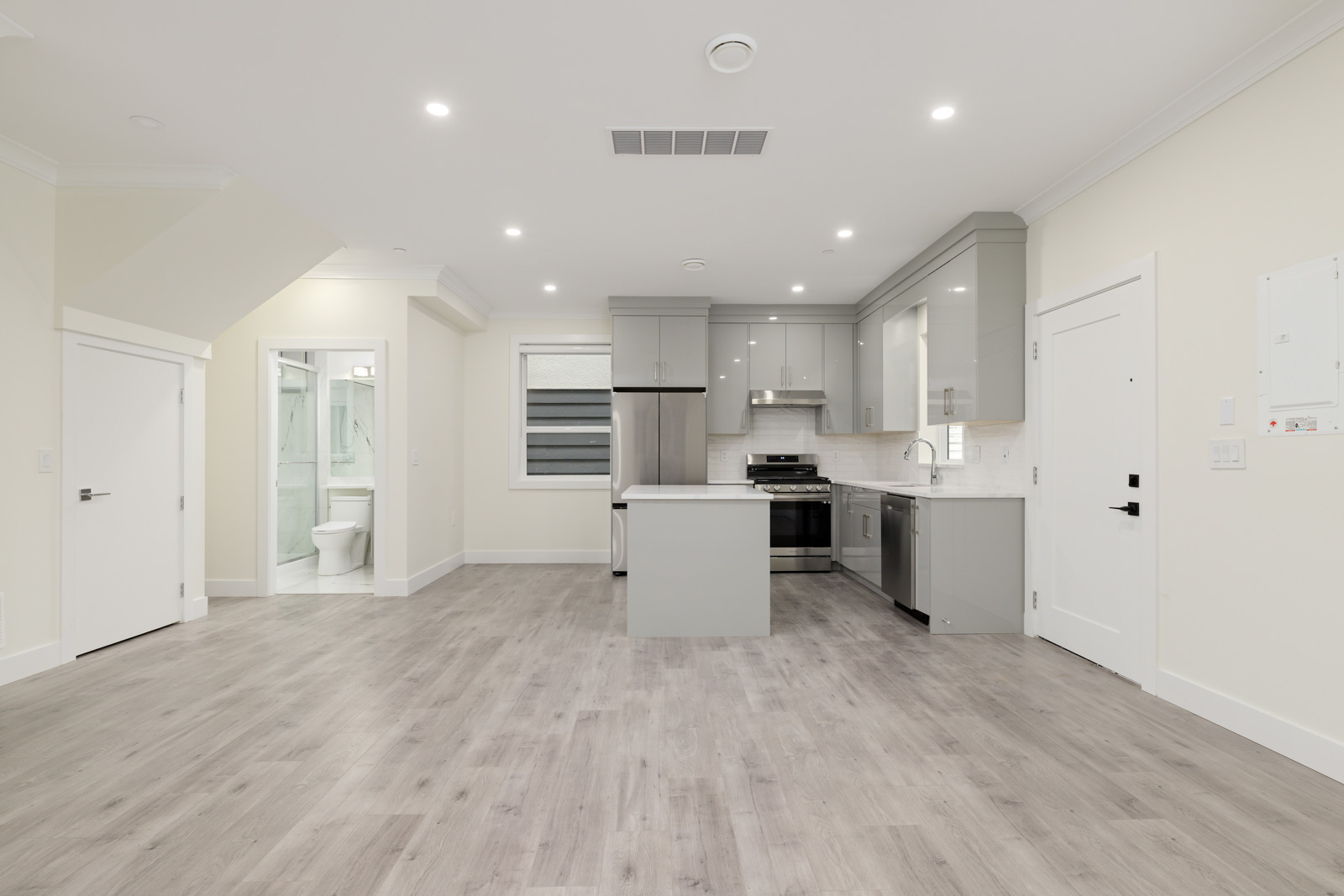 Unfurnished modern kitchen and living area with light wood flooring, grey cabinets, stainless steel appliances, and adjacent bathroom visible through an open door.