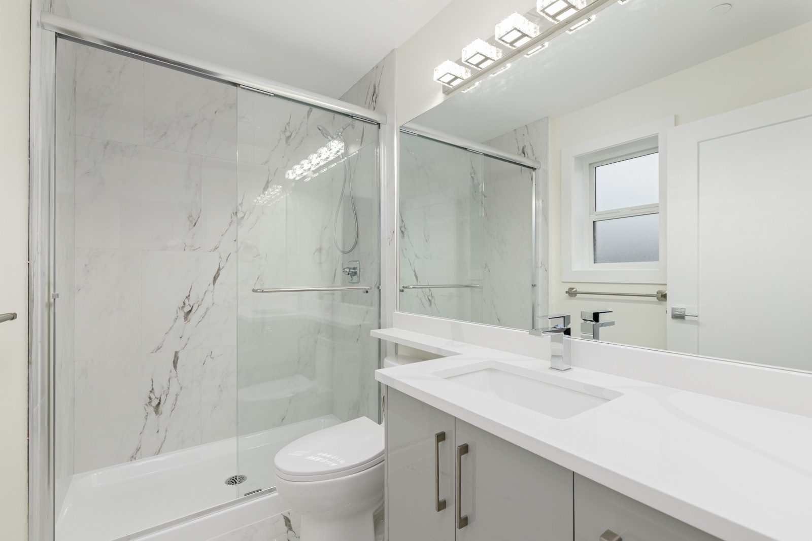 Modern bathroom with a glass-enclosed shower, white marble walls, a toilet, large vanity with sink, and a mirror above the counter.