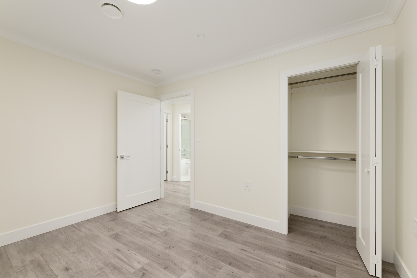 Empty room with light-colored walls, open closet and door, wood-look flooring, and view into a bathroom.