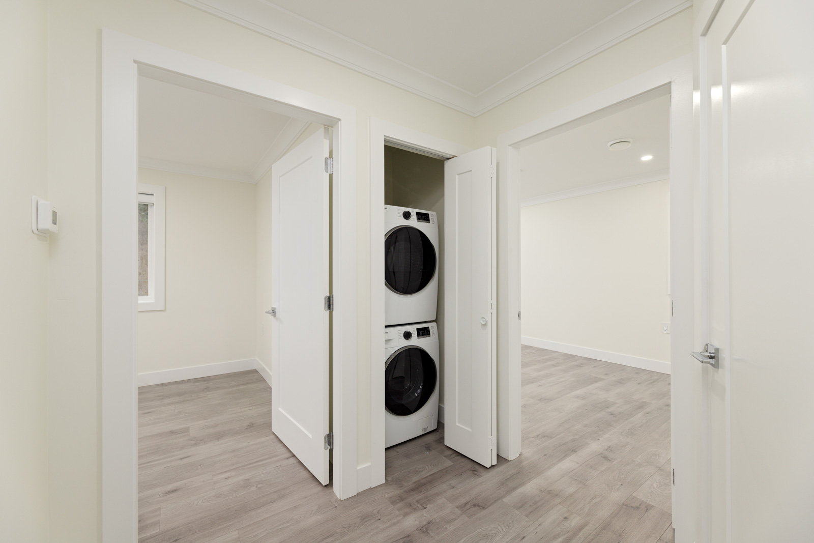 A small laundry closet with a stacked washer and dryer is positioned between two open doors leading to adjacent empty rooms with light-colored walls and wood flooring.