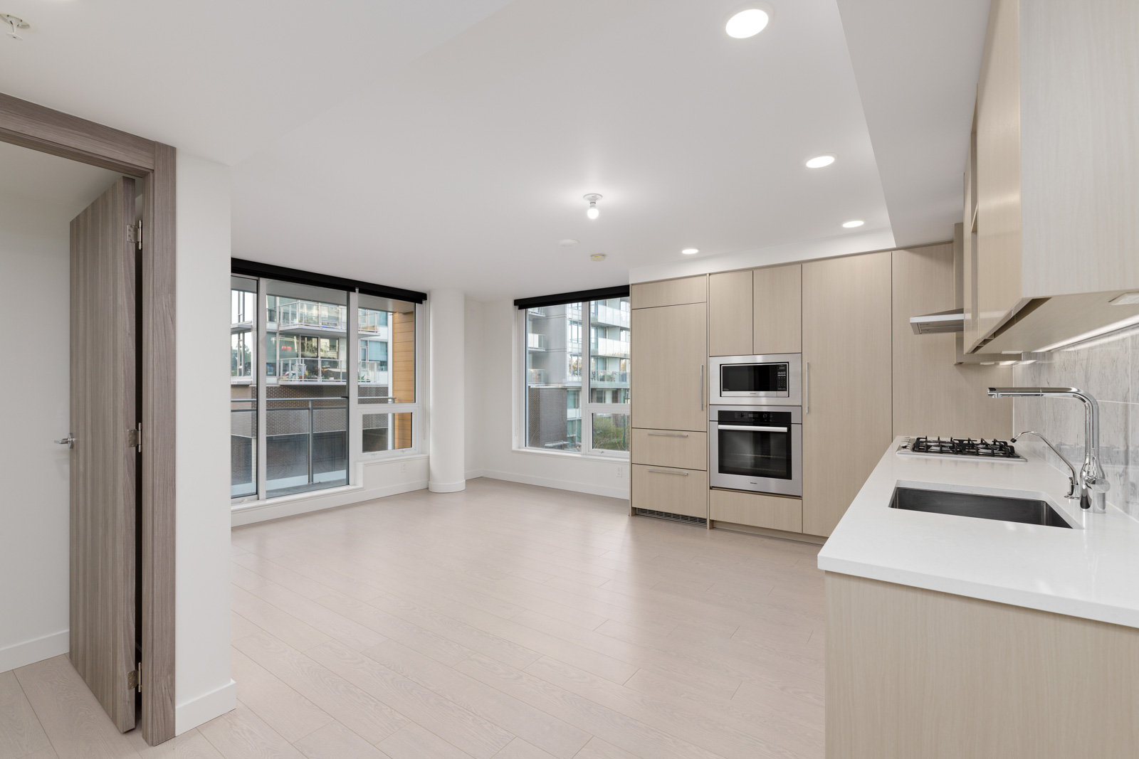 Modern, unfurnished apartment kitchen with light wood cabinets, built-in oven, gas stove, and large windows overlooking neighboring buildings.