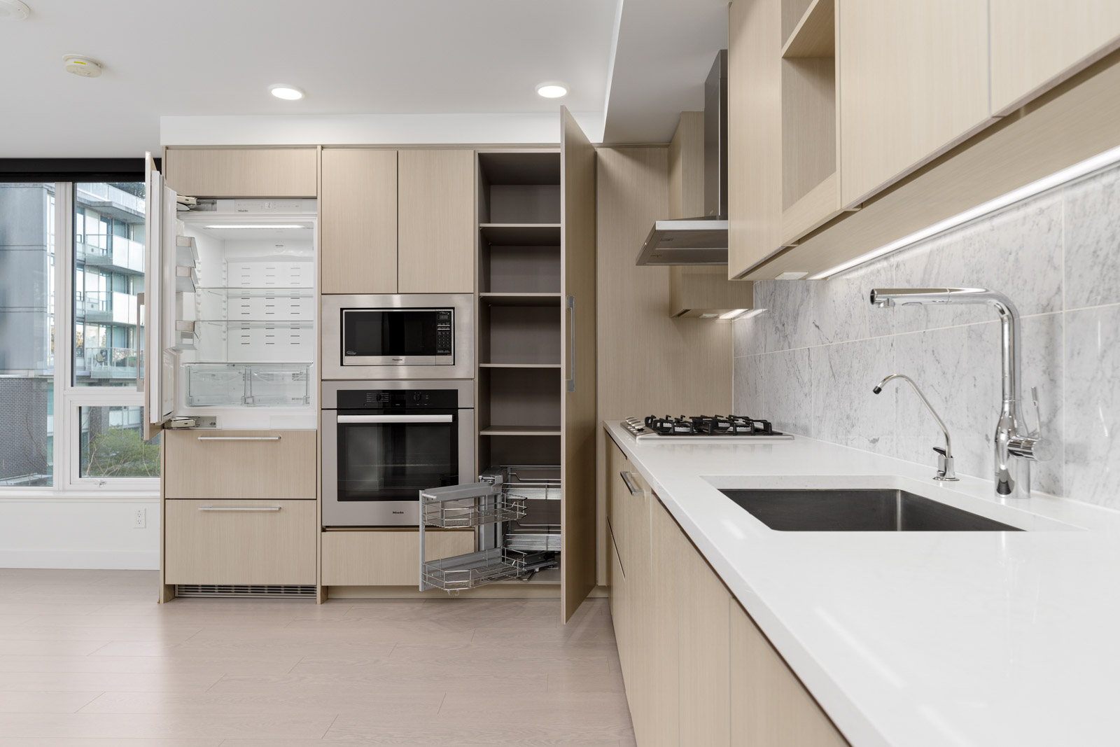Modern kitchen with light wood cabinets, built-in appliances, an open refrigerator, pull-out pantry shelves, gas cooktop, sink, and marble backsplash.