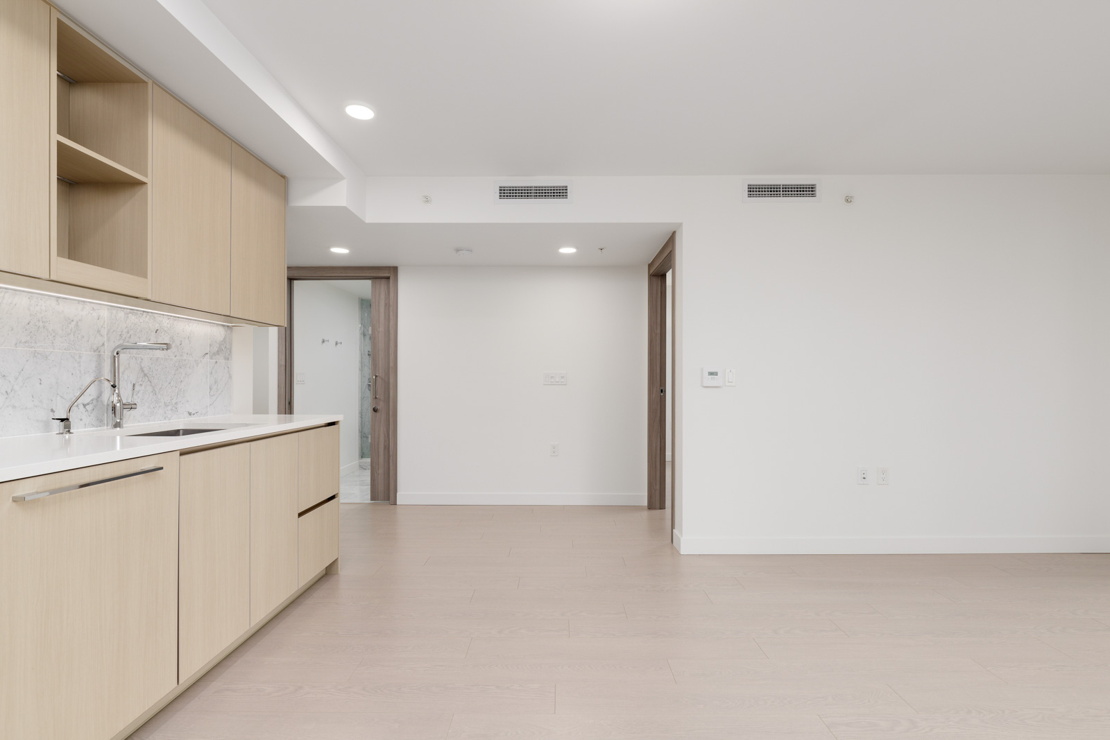A modern, empty kitchen with light wood cabinets, a marble backsplash, and light wood flooring, next to an open space and doorways.