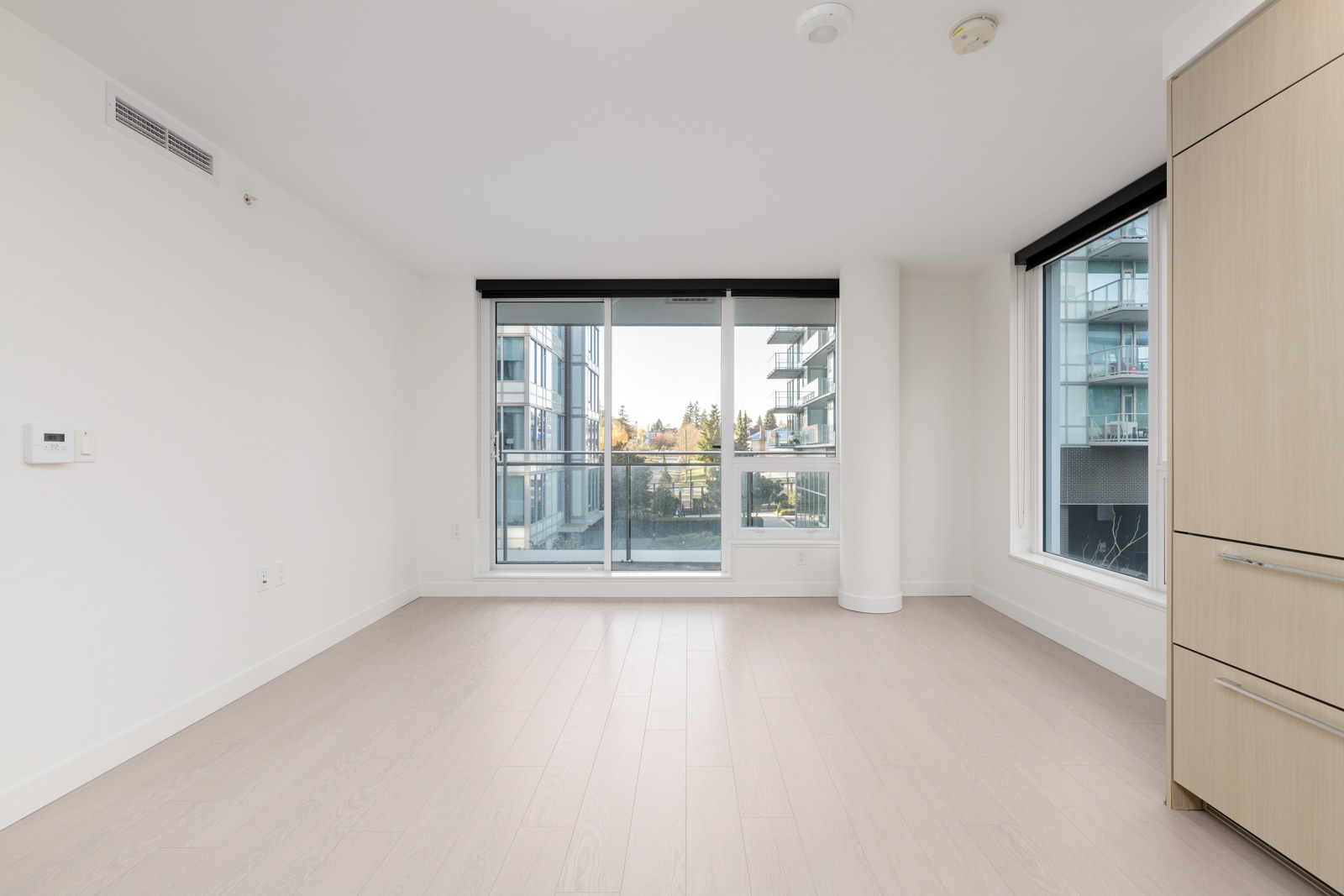 Unfurnished modern apartment room with light wood floors, large windows, and a sliding glass door leading to a balcony with views of neighboring buildings.