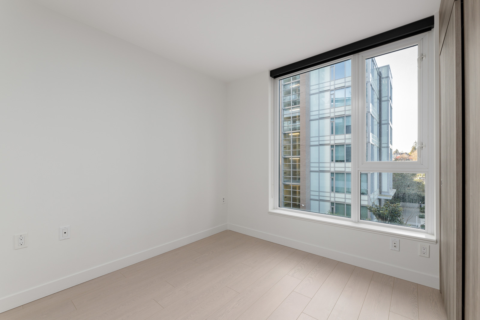 Empty, unfurnished room with light-colored wood floors, white walls, and a large window offering a view of a modern glass building outside.