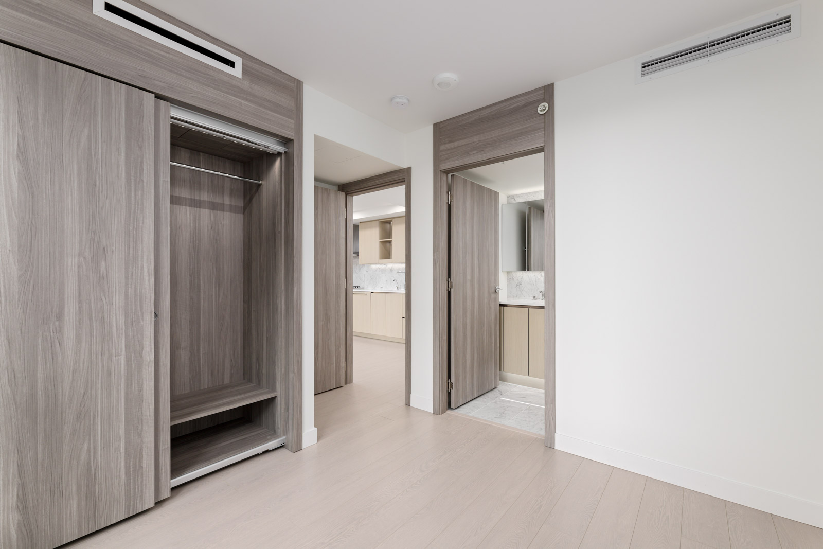 Empty modern room with light wood floors, open closet doors, and open doors leading to a bathroom and another room; neutral color palette throughout.