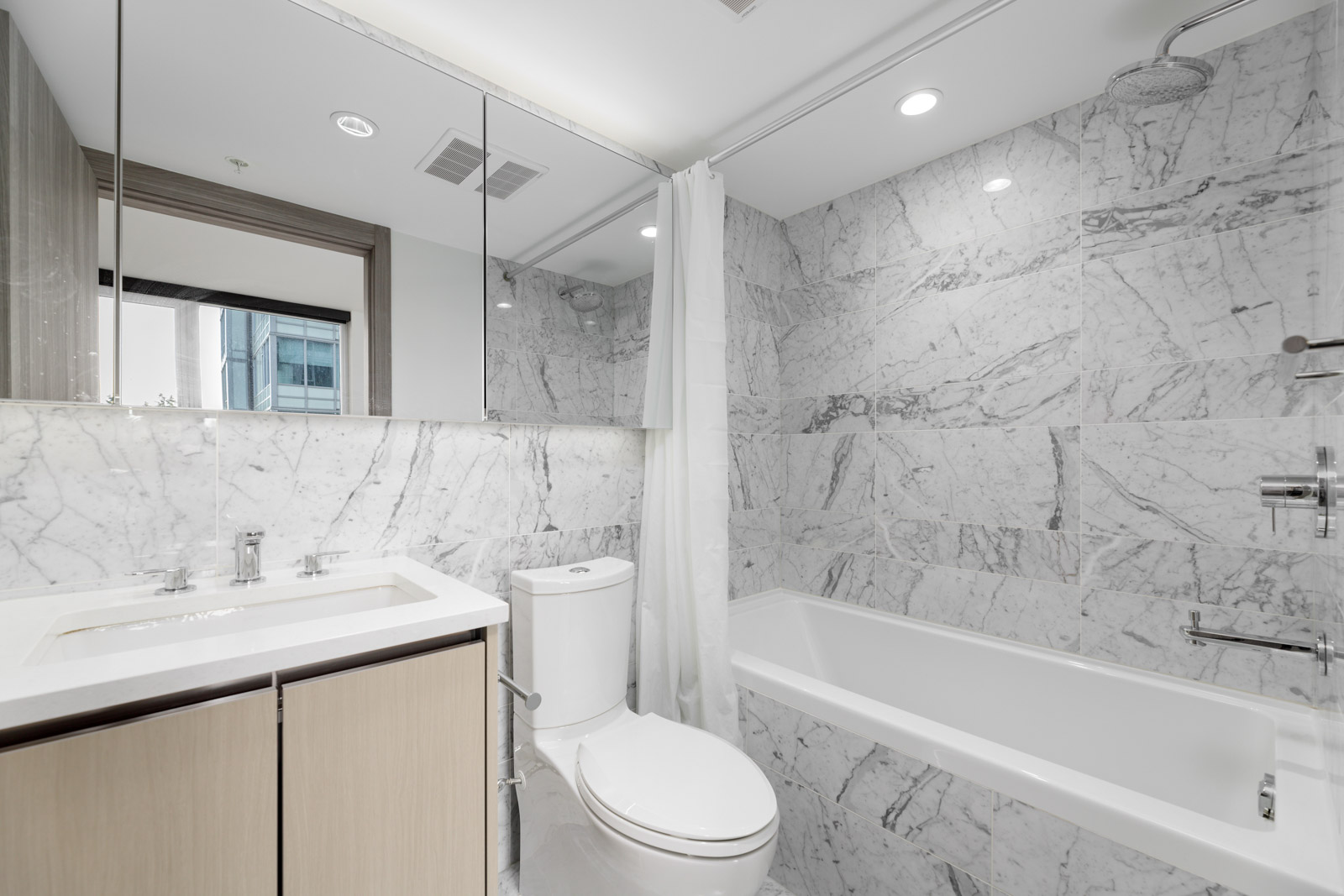 Modern bathroom with marble walls, a white bathtub with shower, a toilet, a sink with wooden cabinet, and a large mirrored wall above the sink.